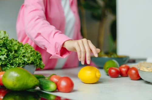 A person in a pink shirt reaching for a lemon surrounded by fresh vegetables.