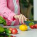 A person in a pink shirt reaching for a lemon surrounded by fresh vegetables.